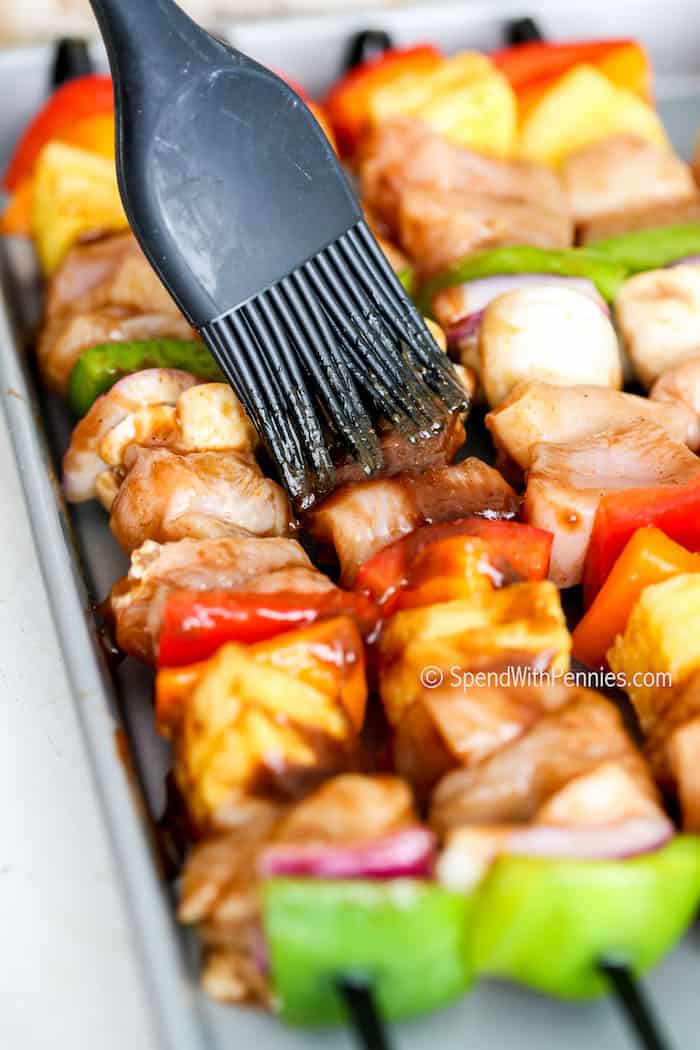 Hawaiian chicken kabobs blend all the tasty flavors of the island. Bite into marinated morsels of juicy tender chicken, an array of vegetables, and sweet juicy pineapple with this easy-to-make recipe. Whether these are made on the BBQ or in the oven, they are the best in summer cooking! #hawaiianchickenkabobs #chickenkabobs #chickenkabob #spendwithpennies brushing sauce on Hawaiian Kabobs before cooking
