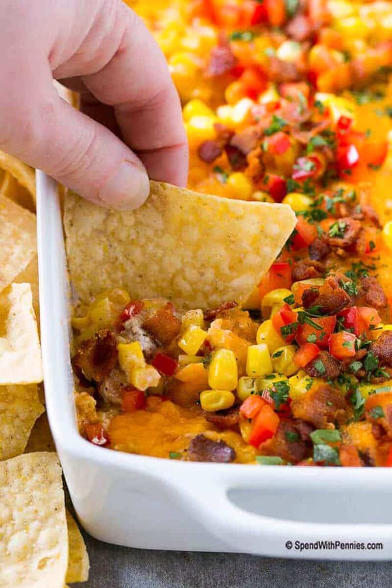 For the best creamy and delicious dip, look no further than this easy corn dip recipe. It's sure to please anyone who tries it, with its thick and creamy cream cheese base, bacon, seasonings, and chunks of sweet corn, peppers, and cheese mixed in. It's simple to make and bake and can be served hot or cold. #corndip #bakedcorndiprecipe #recipecreamcheese #spendwithpennies scooping corn dip from a casserole dish with a chip