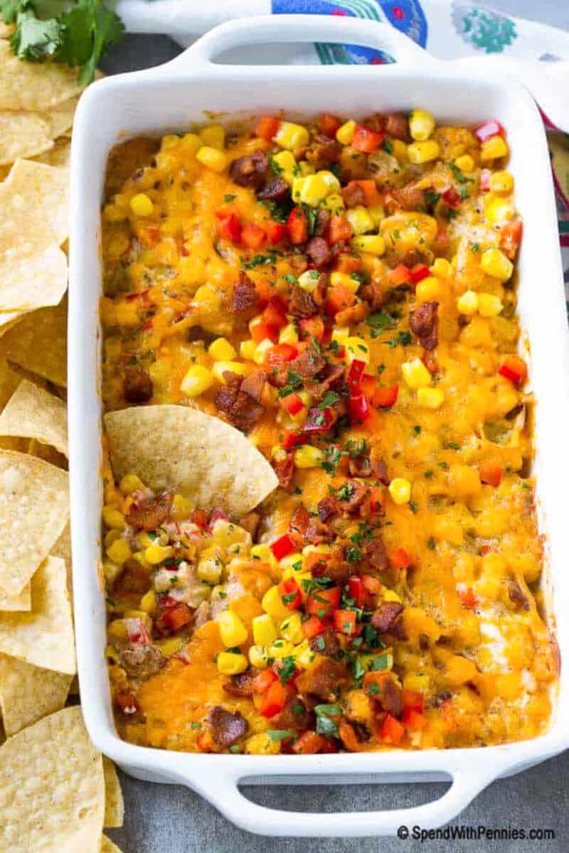 For the best creamy and delicious dip, look no further than this easy corn dip recipe. It's sure to please anyone who tries it, with its thick and creamy cream cheese base, bacon, seasonings, and chunks of sweet corn, peppers, and cheese mixed in. It's simple to make and bake and can be served hot or cold. #corndip #bakedcorndiprecipe #recipecreamcheese #spendwithpennies a chip in corn dip in a casserole dish