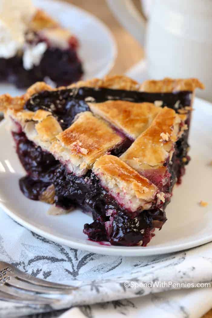 Get ready for a delightful dessert experience with each bite of homemade blueberry pie. Juicy blueberries burst with flavor, perfectly complemented by the buttery crust, creating a truly satisfying treat. #blueberrypie #homemade #pie #dessert #spendwithpennies Slice of Blueberry Pie on a white plate
