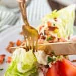 a gold fork and knife cutting into an iceberg wedge topped with bacon, tomato, and blue cheese dressing all on top of a white plate