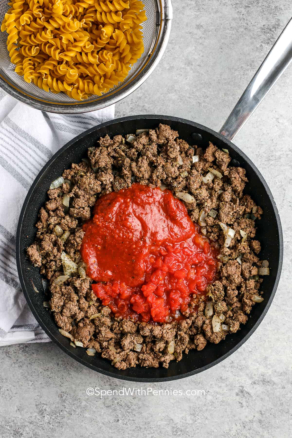 Hamburger casserole is a flexible recipe you can tweak to your liking. Use macaroni, rotini, or other noodles. You could even stir in rice or small diced potatoes to stretch the dish. This bake is loaded with savory tomatoes, beef, and mixed vegetables, then topped with gooey cheese and baked until golden. It’s hearty, flavorful, and truly easy to make. #spendwithpennies tomato sauce with ground beef in a skillet for hamburger casserole