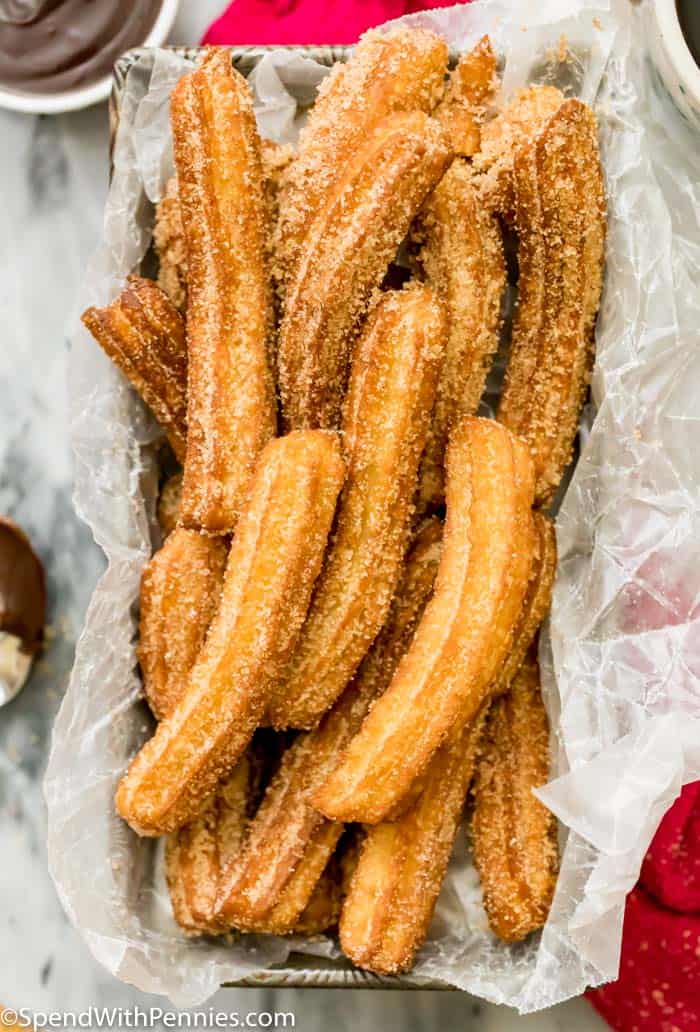 Golden, crispy homemade churros are made from a simple dough, fried until they turn perfectly golden, and coated with a sweet cinnamon-sugar mix. With their delightful crunch and soft, doughy center, homemade churros are a must-try treat for the whole family! #churros #homemade #recipe #mexican #spendwithpennies Churros in a pan with parchment paper