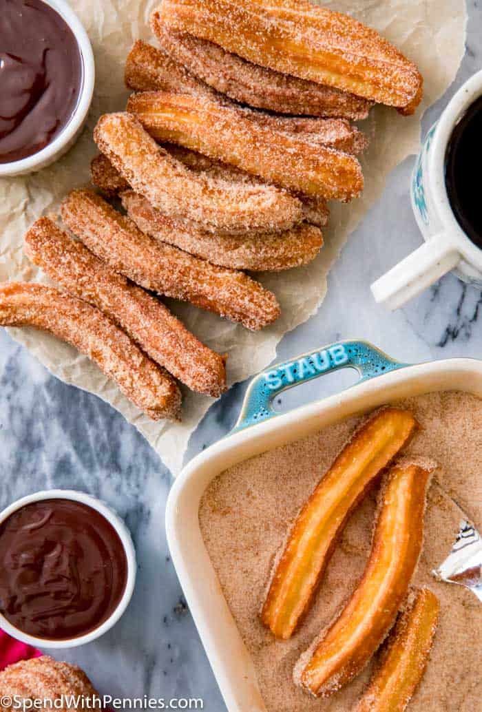 Homemade churros are delicious golden pastries made from a simple dough, fried until they're perfectly golden, and then coated in a sweet cinnamon-sugar mix. These delightful treats have a crunchy outside and a soft, doughy inside, making them a favorite treat for anyone who loves sweets! #churros #homemade #recipe #mexican #spendwithpennies Churros being dipped in sugar