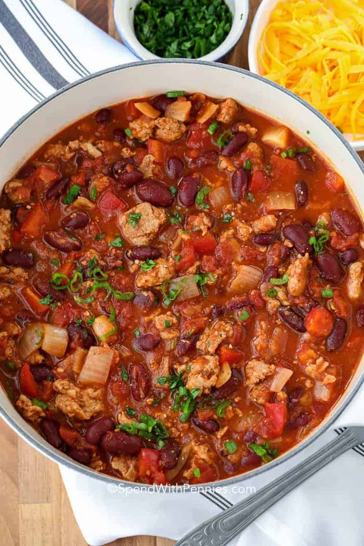 Turkey chili is the ultimate comfort food. In this recipe, ground beef is replaced with ground turkey and then simmered with some tomatoes, kidney beans, broth, and seasonings. Top it with some sour cream, cheese, or guacamole for the best-flavored bowl of chili. This is such an easy and yummy main dish! #turkeychili #turkeychilirecipe #groundturkey #spendwithpennies turkey chili in a pot with a bowl of cheese