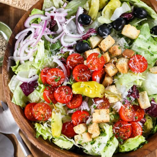 An Italian salad in a wood bowl with dressing and parmesan cheese next to it