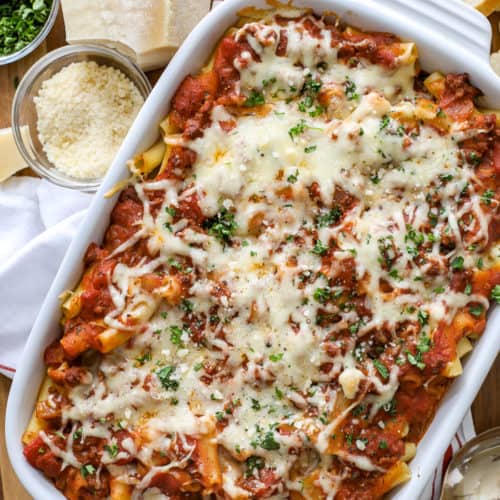 Baked ziti in a white casserole dish with Parmesan cheese and parsley on the side