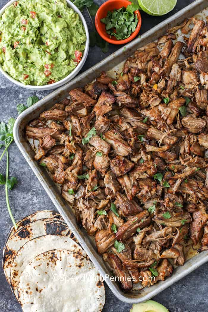 Overhead shot of Pork Carnitas on a baking sheet surrounded by tortillas, limes and avocado