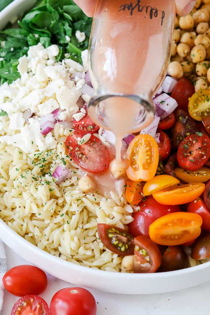 This simple orzo pasta salad is perfect for any summer BBQ or potluck! Made with feta, tomatoes, and garbanzo beans and tossed in a homemade lemony dressing, it is a definite crowd-pleaser. Everyone loves this colorful and flavorful pasta salad! #orzopastasalad #pastasalad #sidedish #spendwithpennies Pouring dressing over Orzo pasta salad with tomatoes, spinach and chickpeas in a bowl