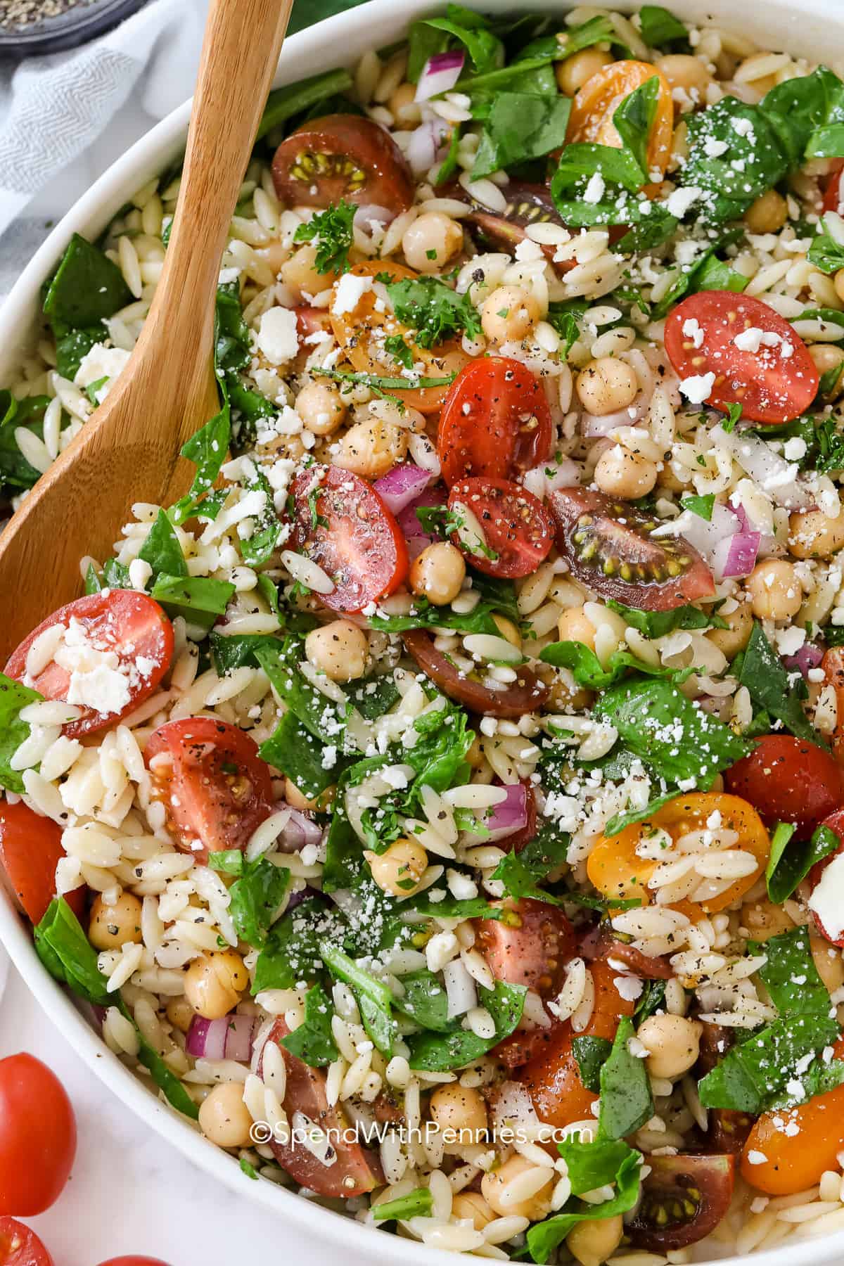 This simple orzo spinach pasta salad is a summertime favorite! Made with feta, tomatoes, and garbanzo beans tossed in a homemade lemony dressing, it's perfect for a BBQ or potluck. Serve cold at your next barbecue! #orzopastasalad #pastasalad #sidedish #spendwithpennies serving orzo pasta salad with a wooden spoon