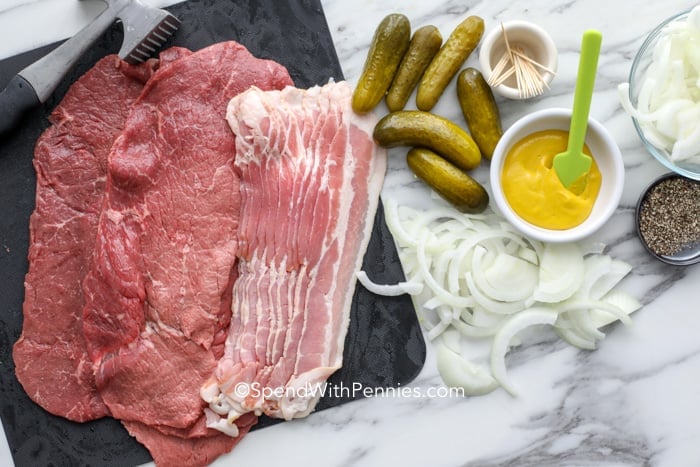 Make this easy Rouladen recipe in the oven or in the slow cooker. Start with thinly sliced inside round or flank steak and cover it in mustard, onions, bacon and wrap around a dill pickle! This classic German recipe is so delicious and well worth the wait! #spendwithpennies #rouladen #maincourse #beefrecipe Ingredients for beef rouladen on a marble board