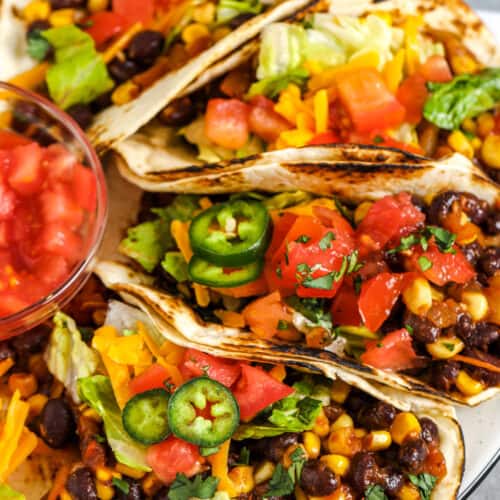 Four black bean tacos prepared on a plate. Topped with lettuce, tomatoes, cheese, and jalapenos.