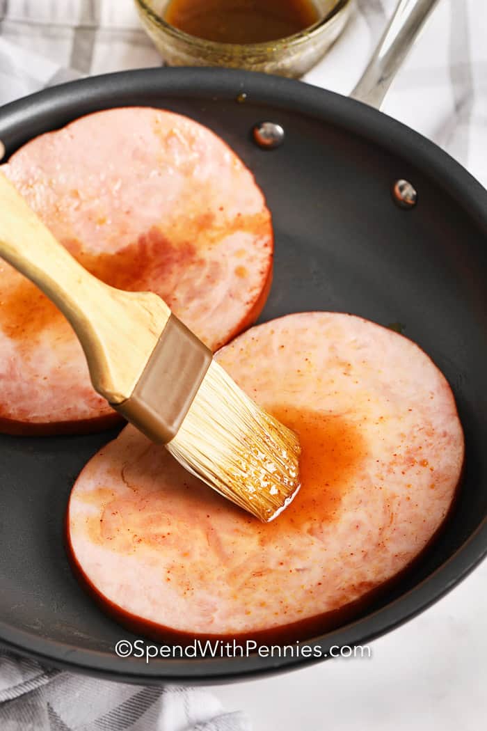 Savory ham steak is fried with a deliciously sweet glaze, then served with mustard on the side. Make this recipe for breakfast, lunch or dinner! It goes great with any side dish like roasted or mashed potatoes, sautéed veggies, or a side salad. Don’t forget the garlic bread! #glazedhamsteak #maindish #hamsteak #spendwithpennies Ham Steak in a pan being glazed