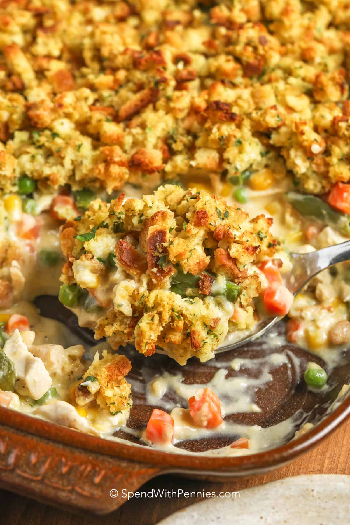 Easy chicken stuffing casserole turns rotisserie chicken into a warm, satisfying dinner with very little prep. Cream of chicken soup and sour cream make the filling rich and creamy, while mixed vegetables add color, and stuffing mix bakes into a savory golden topping. This simple baked casserole is ideal for chilly evenings, leftover chicken, and busy family dinners. Save this chicken casserole with stuffing for an easy weeknight meal. #spendwithpennies A spoon in a chicken and rice casserole in a brown dish.