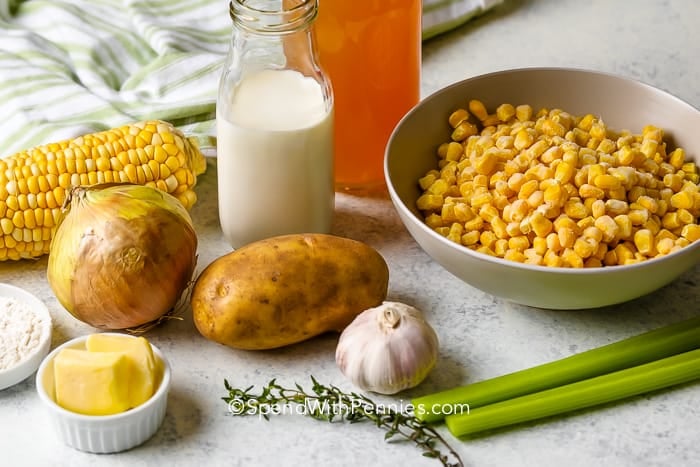 Corn kernel in a bowl with a corn on the cob, an onion, a potato, clove of garlic, jar of cream, side of butter, celery stalks, tablespoon of flour, and chives