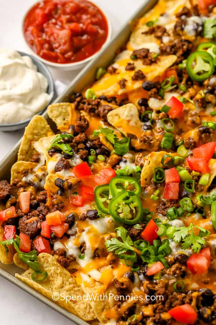 A tray full of baked nachos topped with ground beef, black beans, green onions, tomatoes, jalapenos and shredded cheese, with a side of salsa and sour cream