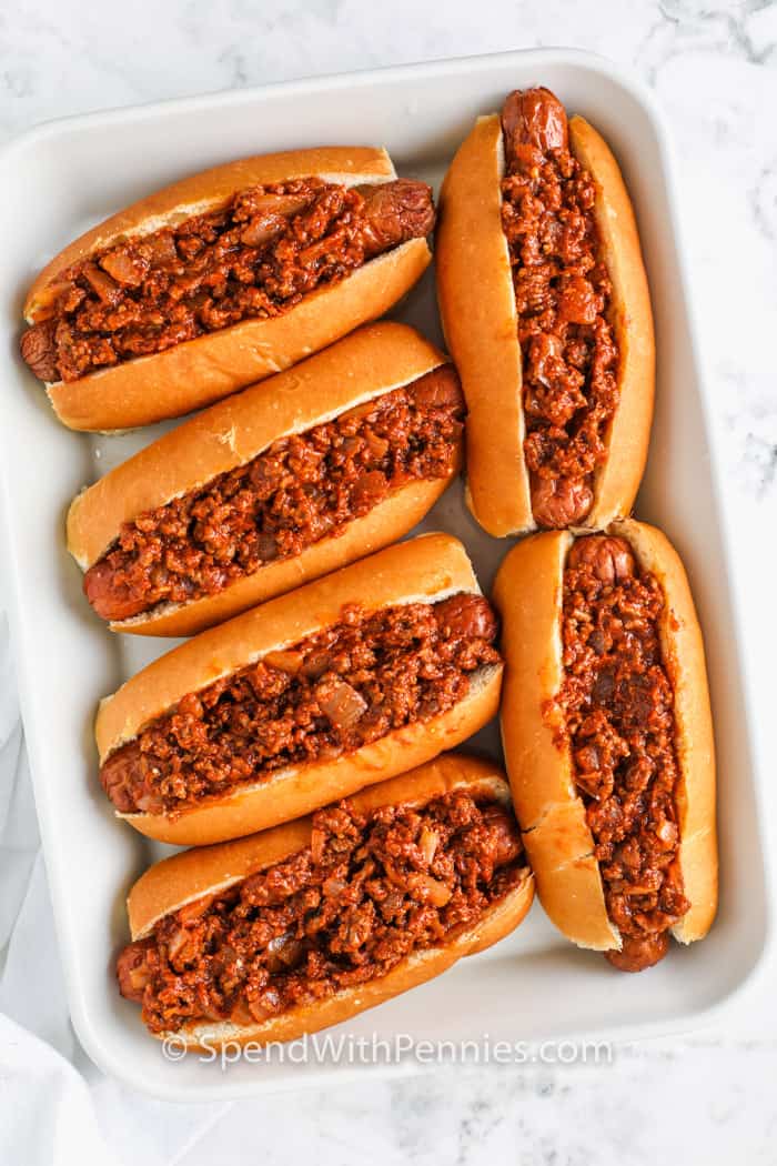 Chili cheese dogs are a hot recipe that always hits the spot whether it's game day, a movie night, or just because. Juicy hot dogs are baked in buttery top-cut buns, loaded with savory chili, and finished with a gooey blanket of cheddar. This is the best kind of comfort food—simple, hearty, and totally satisfying. Try turning leftovers into a chili cheese dog casserole for an easy next-day meal. Make a full tray and keep it warm in the oven for serving a crowd. #spendwithpennies adding chili to bun to make Chili Cheese Dogs