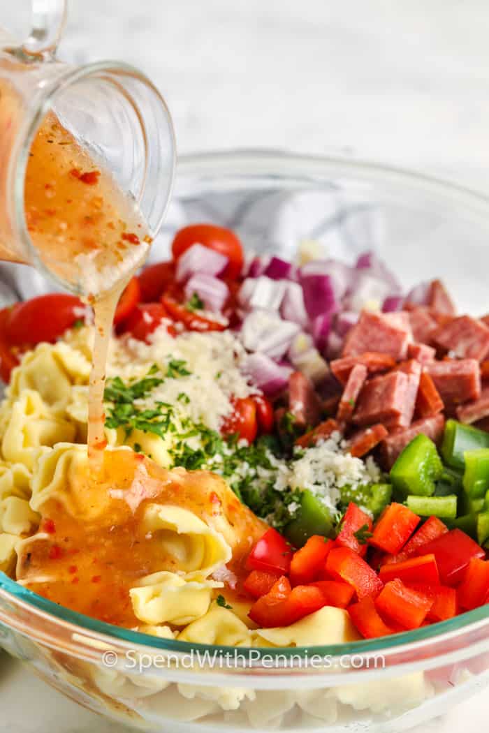 This bright & colorful Italian Tortellini Salad is the perfect dish for a potluck or party! It's loaded with fresh veggies, tortellini, meat, & cheese! #italiantortellinisalad #tortellinisalad #pastasalad #spendwithpennies pouring dressing over top ingredients to make Italian Tortellini Salad