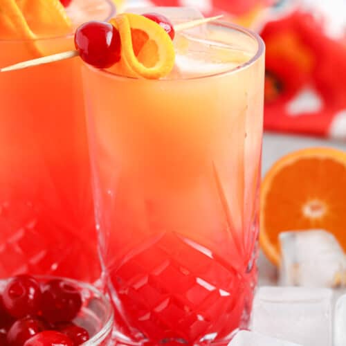 two glasses of Tequila Sunrise with cherries , oranges and ice cubes