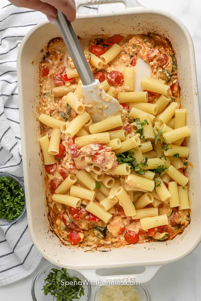 mixing cooked pasta and parsley with cooked cherry tomatoes, chopped zucchini, red pepper, onion, and feta cheese in pan