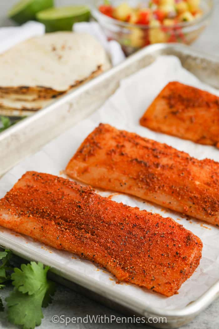 Mahi Mahi Tacos can be baked in the oven or cooked on the grill. The seasoned fish is shredded, wrapped in a corn tortilla, and topped with some cabbage or salsa. #spendwithpennies fish on a baking sheet with ingredients in the back to make Mahi Mahi Tacos
