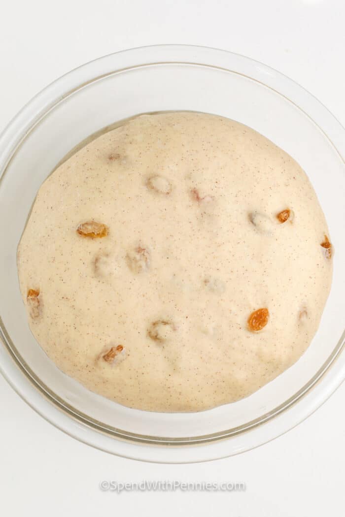 Begin your day on a delicious note with a batch of flavorful hot cross buns. These soft buns are enriched with orange zest, nutmeg, cinnamon, and sweet raisins. Pair them with a comforting cup of hot chocolate for a cozy morning indulgence. And make the most of any leftovers by transforming them into a delightful bread pudding! #hotcrossbus #hotcrossbunsrecipe #spendwithpennies #hotcrossbunsrecipeeasy Hot Cross Buns dough rising in a bowl