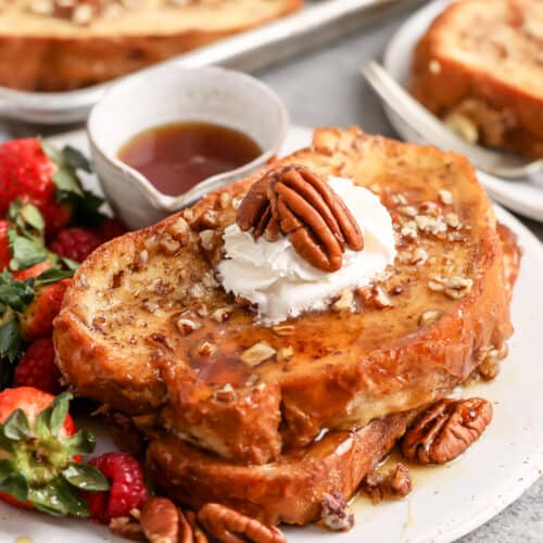 Two slices of overnight french toast being served topped with whipped cream and pecans