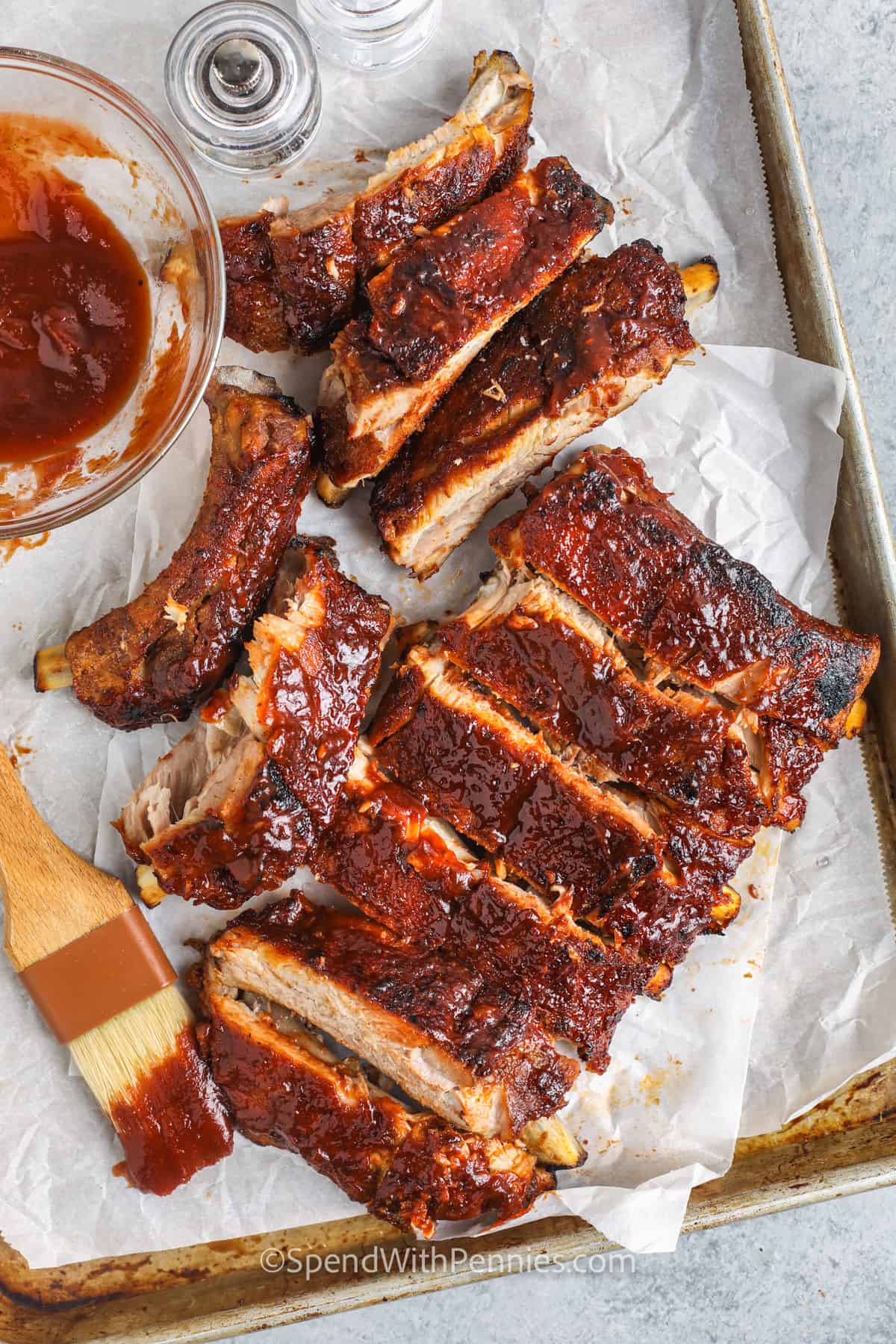Tender oven-baked ribs are so juicy and tender that they fall right off the bone. Seasoned with a homemade rib rub and smothered in your favorite BBQ sauce, they are packed with flavor! Seal with a quick grill and enjoy these finger-licking good ribs any time of the year. #ovenbakedribs #ribs #recipe #spendwithpennies cut Oven Baked Ribs on a sheet pan with sauce