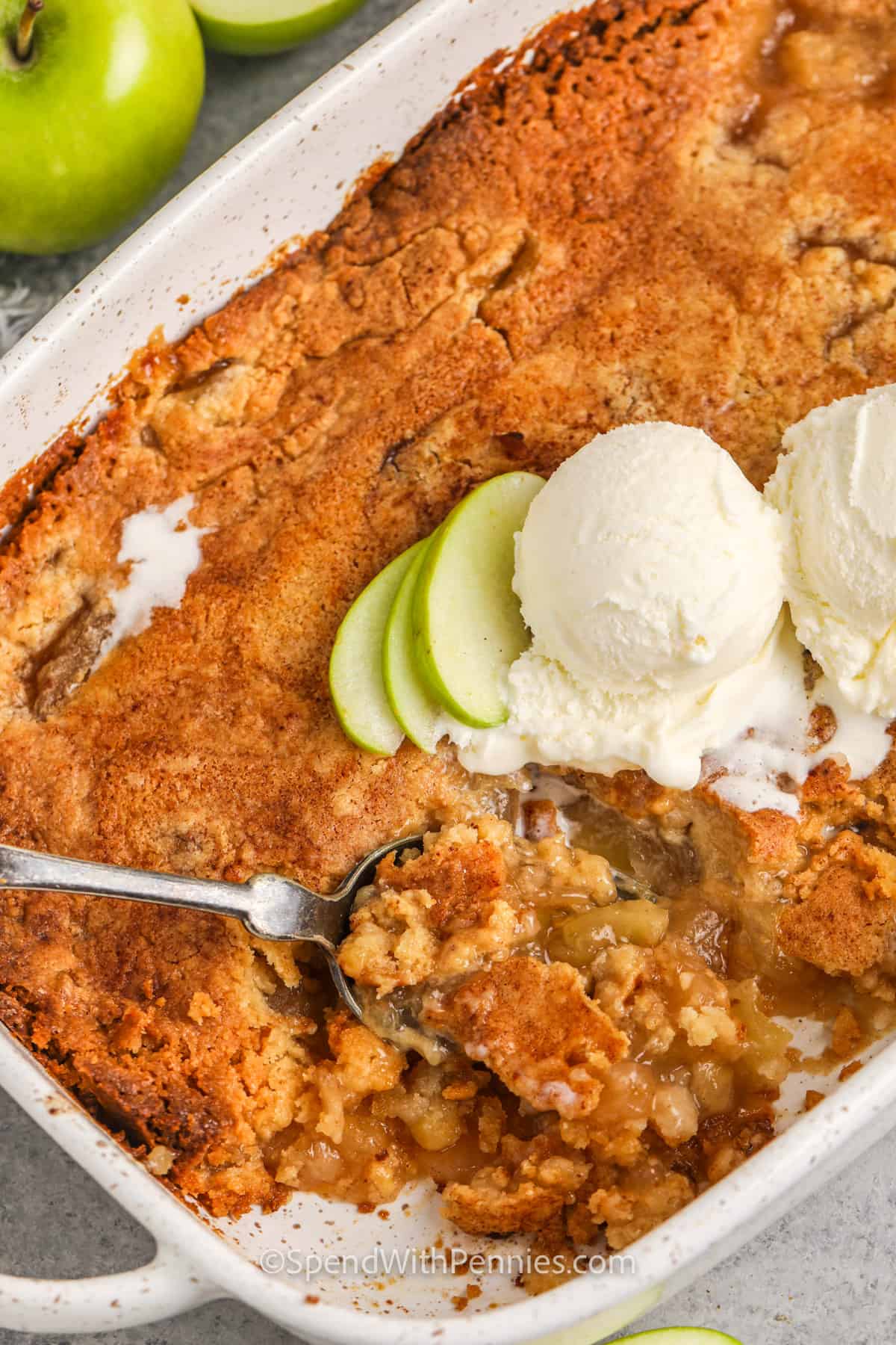 Cook a sweet and easy apple dump cake for dessert. There are only 4 ingredients in this recipe - cake mix, butter, apple pie filling, and some cinnamon. Just mix them together, add them to a pan, and cook. What could be more simple than that? Top it with vanilla ice cream, or some caramel sauce for the grand finale. #appledumpcake #dumpcakewithapples #dumpcake #spendwithpennies cooked Apple Dump Cake in the pan with a spoon