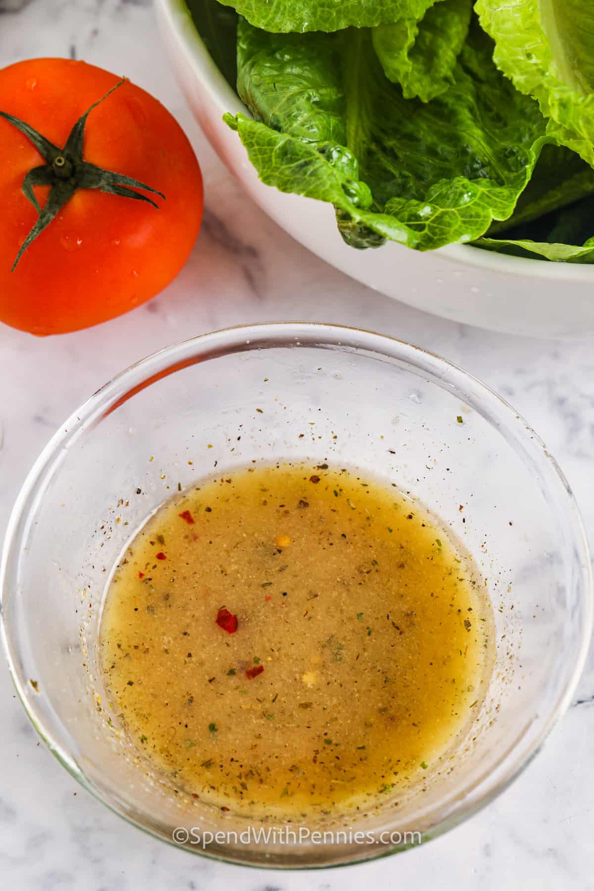 Denna syrliga och lite söta italienska dressing är balanserad och redo att användas på pastasallader, eller som en marinad för kyckling eller nötkött. Hemlagad salladsdressing smakar alltid bäst, och det här receptet kommer säkerligen att bli en häftklammer i köket med sina enkla ingredienser och ljusa smak. Prova den på en trädgårdssallad eller använd den som vitlöksbrödsdipp! #italienskdressing #hemlagad marinad #dressingrecept #spendwithpennies Italiensk dressing i en skål med sallad och tomat på sidan
