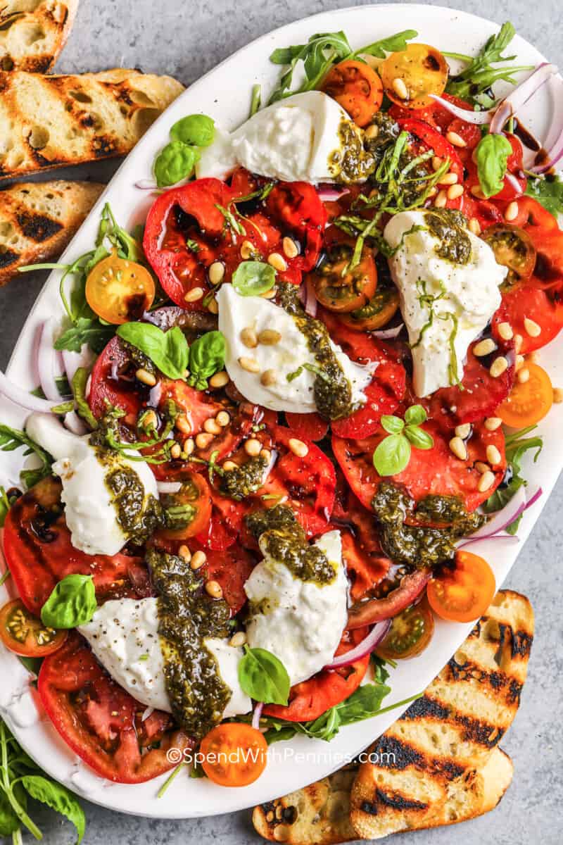 Everyone will love this burrata salad. Try a new style of garden salad made with a tangy balsamic glaze and fresh burrata cheese. This salad can be made with peach and tomato, beet and arugula, or stick with the recipe and use basil and pesto. Any way you serve it up it will get rave reviews! #burratasalad #arugula #tomato #spendwithpennies tomato burrata salad on a large platter