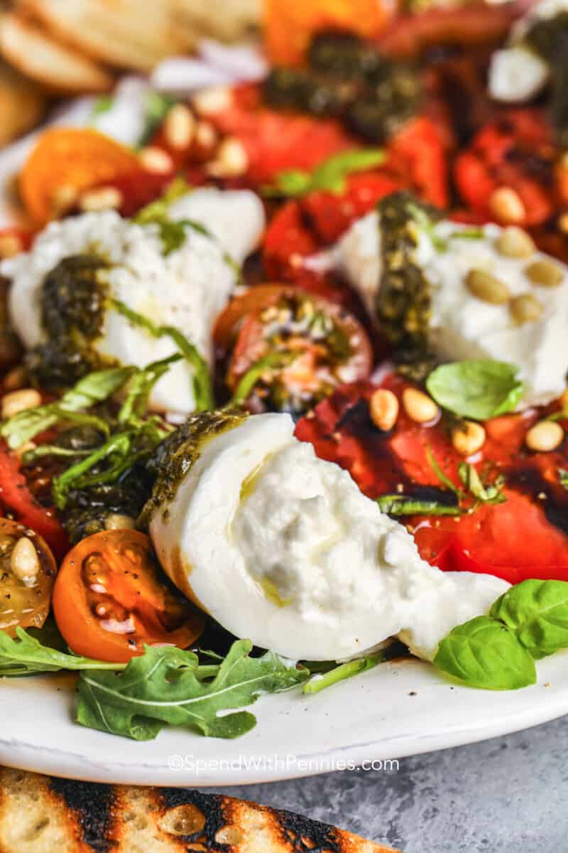 Make an excellent burrata salad with this easy-to-make recipe. Garden tomatoes with peaches or watermelon make a great variation. Or have it with beets and burrata. Just slice and toss with a tangy balsamic glaze and some basil, if desired. Serve with pine nuts on top and add some pesto too! It is so delicious. #burratasalad #arugula #tomato #spendwithpennies close up of burrata on a salad