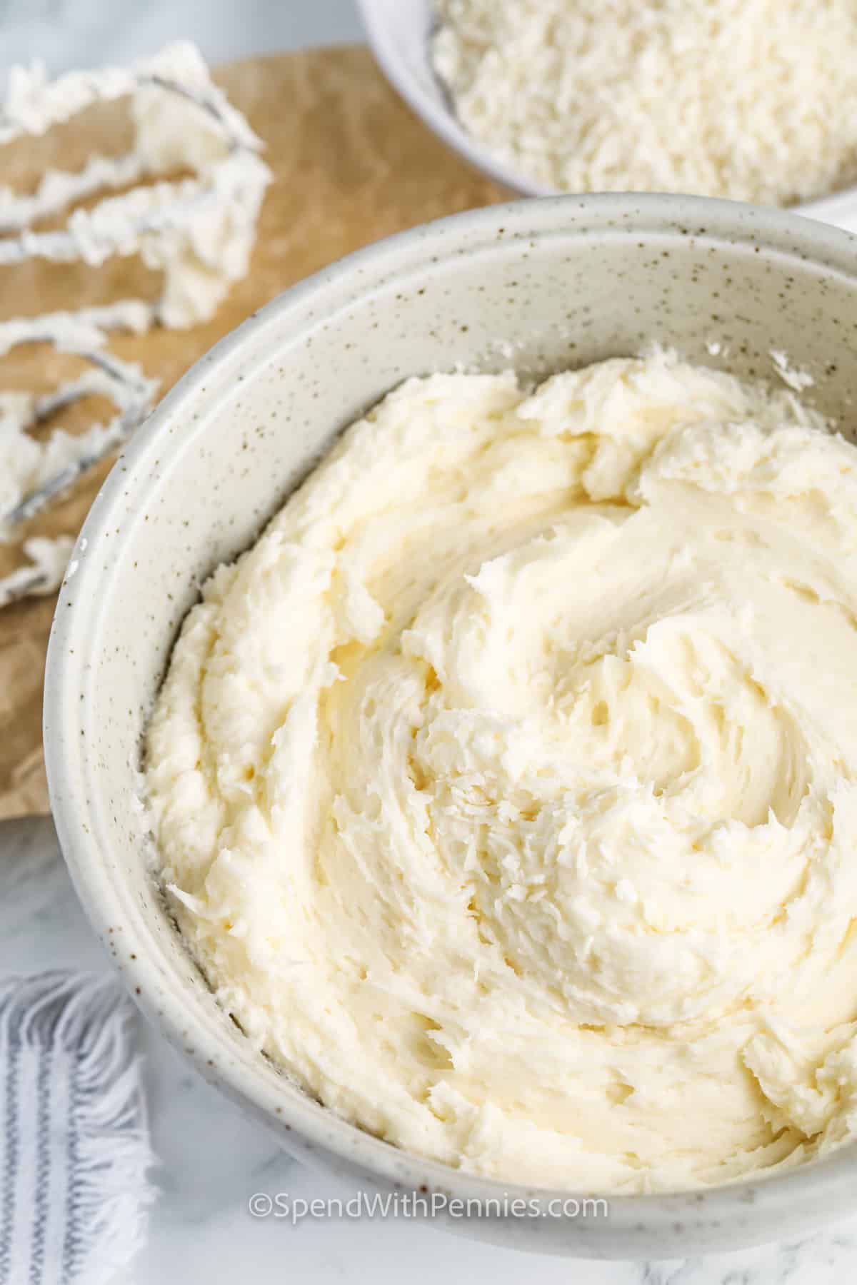 A layer cake covered with swirls of coconut frosting is the perfect summer birthday treat! This easy recipe is a tasty variation of classic buttercream and takes only minutes to make. Just whip butter until fluffy and light, then fold in powdered sugar, coconut flavoring, and coconut cream. Then spread or pipe it onto a layer cake or cupcakes. Sprinkle with chopped pecans or toasted coconut! #coconutfrosting #spendwithpennies #coconutfrostingrecipe #coconutfrostingeasy bowl of Coconut Frosting with a whisk beside it