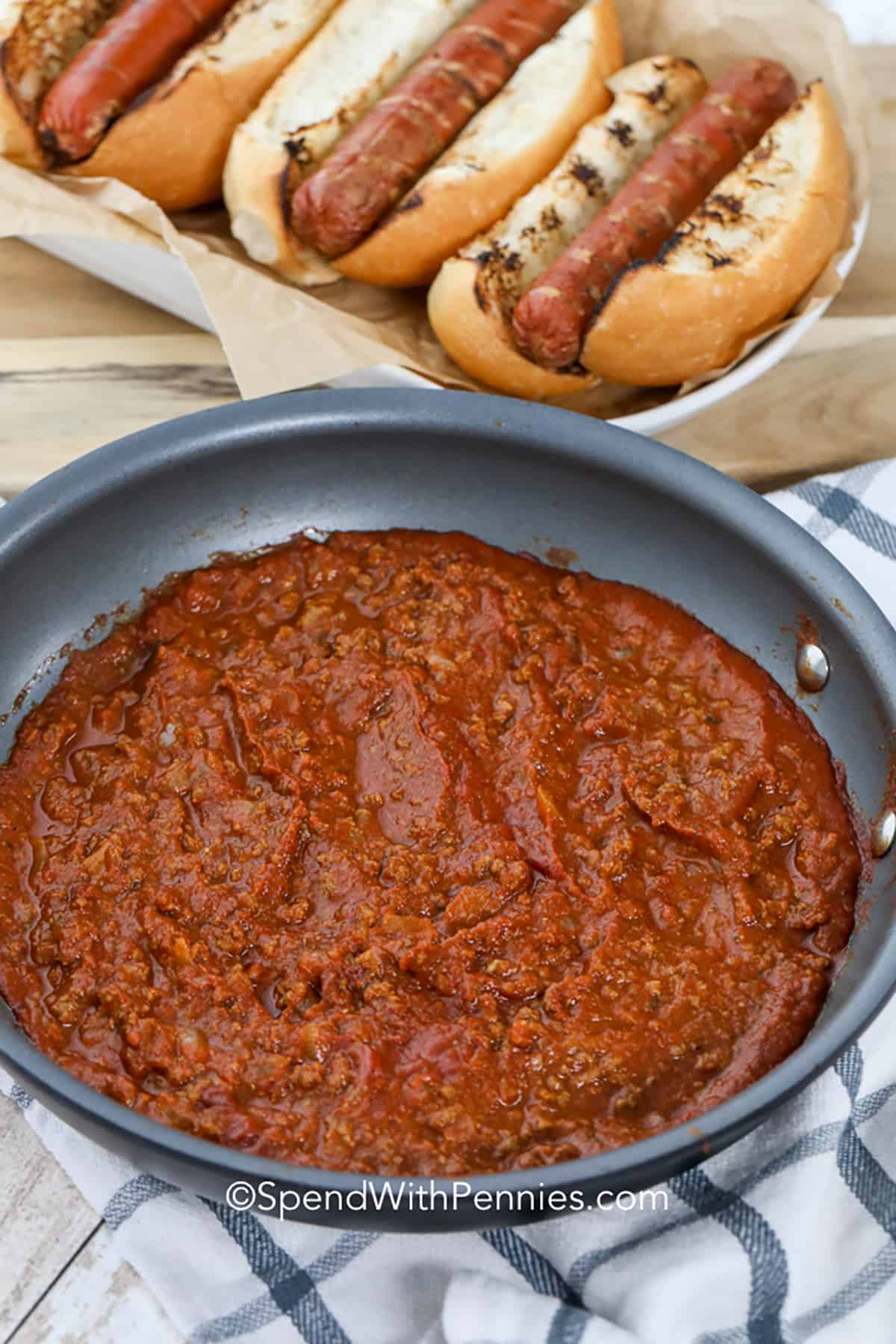 To enjoy this chili dog sauce at its finest, generously spoon it over your hot dog and garnish with chopped pickles and cheese. It's an effortless recipe that you can prepare in no time or store in the freezer for future use. With just ground beef, tomato sauce, and a blend of seasonings, you'll have a versatile and delightful condiment for any occasion! #chilisauce #hotdogchilisauce #condiment #hotdogchili #spendwithpennies Quick Hot Dog Chili in the pan with hot dogs in the back