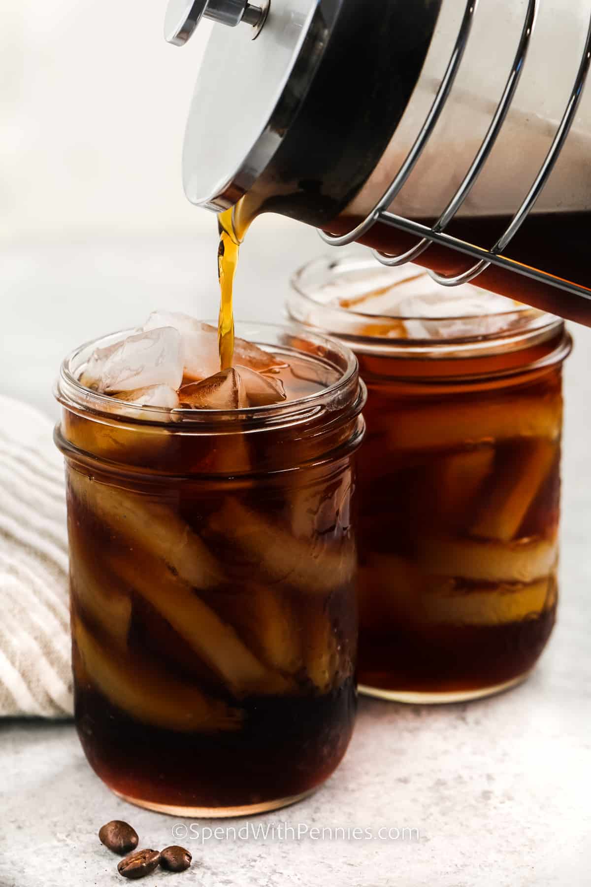 Enjoy a coffee cold brew at home with this easy-to-make recipe. Why head out to the local Starbucks and Dunkin Donuts for a cold brew, when it can be made at home? To make this recipe, simply let the coffee soak with water in a French press for a couple of hours. The coffee can then be topped with some whipped cream, or pumpkin spice for extra flavor. #coldbrew #coldbrewcoffee #coldbrewrecipe #spendwithpennies pouring Cold Brew in glasses of ice