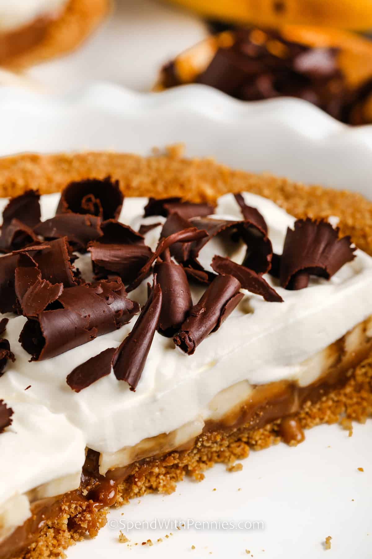 A decadent dessert of banoffee pie is just what is needed to satisfy any sweet tooth. It all begins with a graham cracker crust that is layered with sweet caramel and bananas and then topped with smooth and creamy whipped cream. Garnish with some chocolate curls for a dessert that they won't forget. #banoffeepie #banoffeepierecipe #banoffeedessert #spendwithpennies half of a Banoffee Pie in a white dish