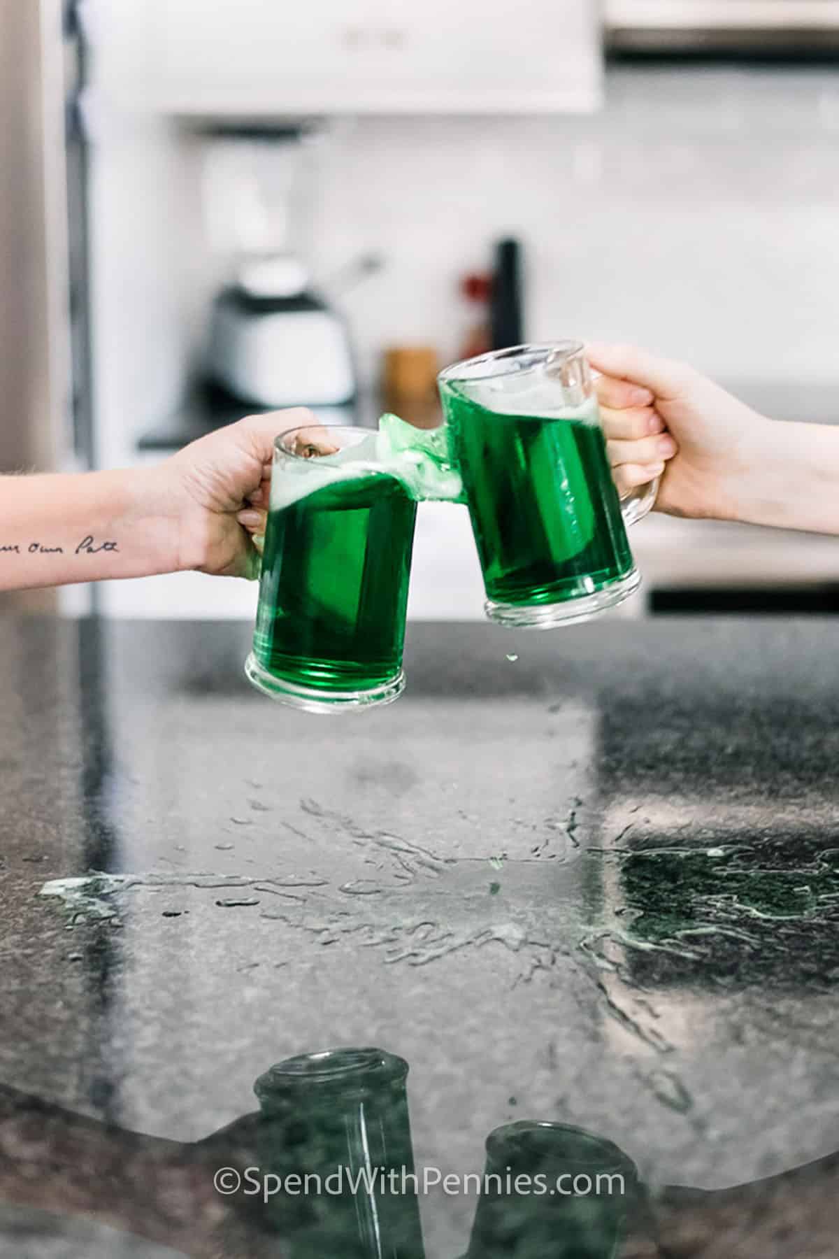 two hands toasting with glasses of green beer, with beer spilling out onto counter