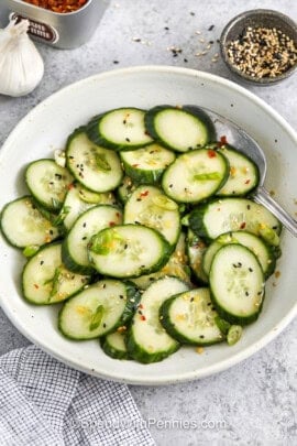 asian cucumber salad in a white bowl with a serving spoon, garlic and a small bowl of spices