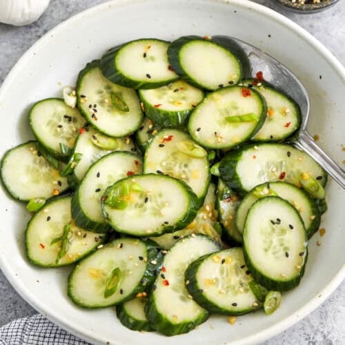 asian cucumber salad in a white bowl with a serving spoon, garlic and a small bowl of spices