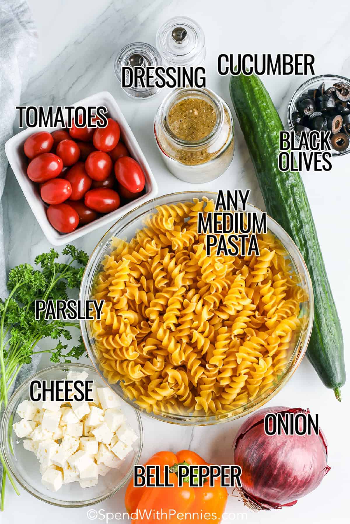 ingredients for easy pasta salad including dressing, cucumber, pasta, tomatoes, cheese, bell peppers, & onion, black olives and parsley
