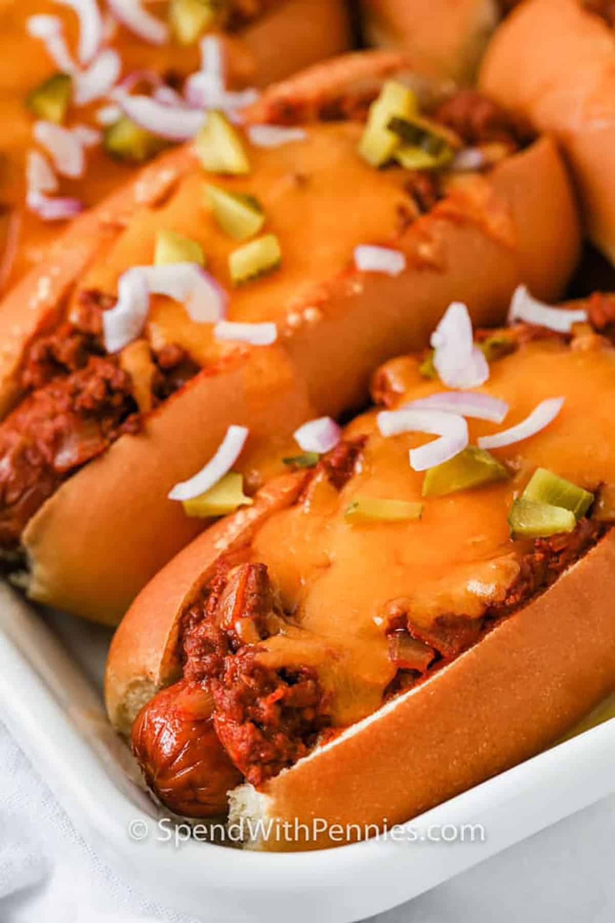 Chili cheese dogs take everything you love about comfort food and bake it into one delicious dish. Toasted buns, warm chili, and melted cheese make this hot recipe the best pick for parties, sleepovers, or movie nights. Make it simple with canned chili or go homemade for that extra touch. Or layer it up casserole-style for a fun twist on the original. Don’t forget a sprinkle of green onions or jalapenos before serving. #spendwithpennies close up of Chili Cheese Dogs on a plate