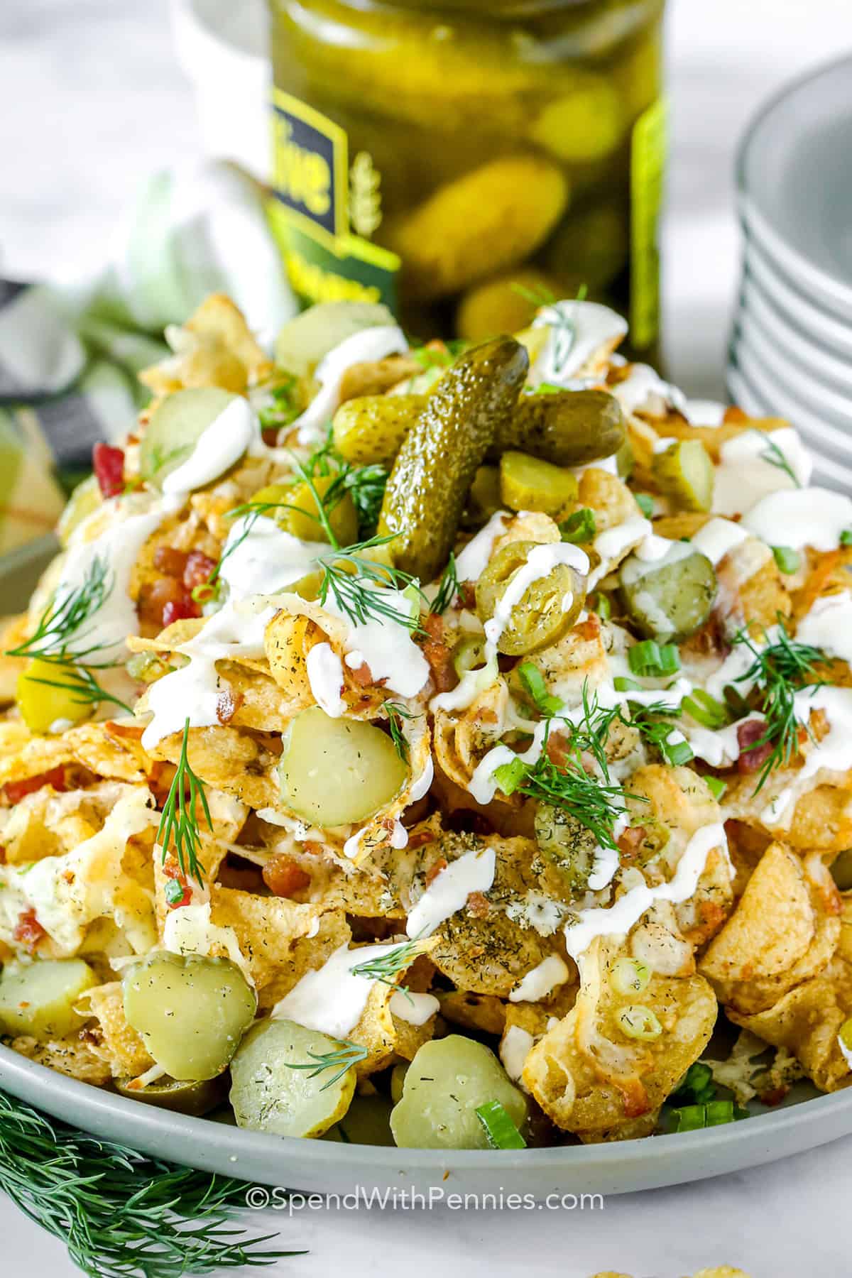 Dill pickle nachos are a must-try for anyone who loves the bold flavor of pickles in a new way. With crunchy kettle chips, cheese, bacon, and pickles layered together, each bite is loaded with flavor. Sour cream dipping sauce adds a cool, creamy finish. #spendwithpennies cropped dill pickle nachos on a platter drizzled with sour cream sauce.