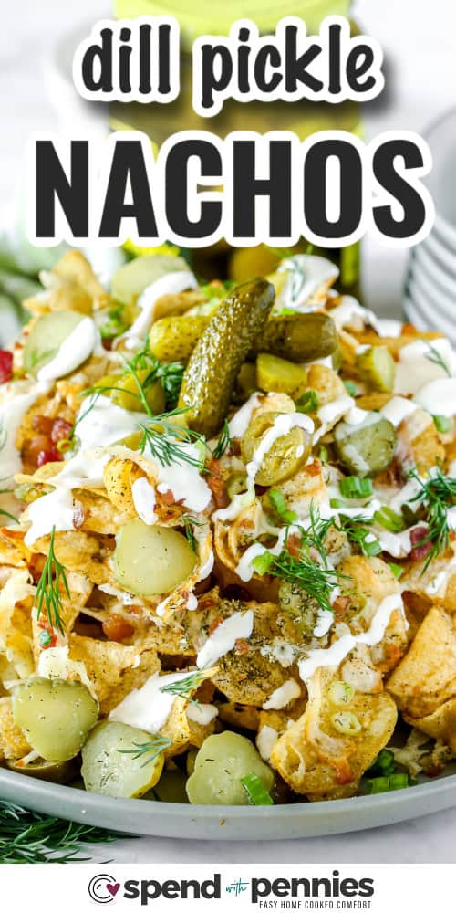 Dill pickle nachos are layered with crispy chips, gooey cheese, and plenty of tangy pickles for a unique twist. Topped with bacon and green onions, they bake up bubbly and golden in just minutes. Perfect with a cool, creamy sour cream dipping sauce for extra flavor. #spendwithpennies dill pickle nachos on a plate with text