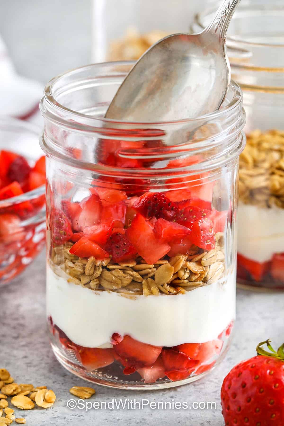 This strawberry parfait is creamy, fruity, and crunchy all in one easy-to-make recipe. Just layer yogurt, strawberries, and granola in a cup or jar for a quick treat. Mix things up with blueberries, jello, or even a bit of chocolate. It’s light in calories and can turn into a shortcake-style dessert with a swirl of cheesecake filling or whipped cream. #spendwithpennies adding strawberries to jar to make Strawberry Parfait