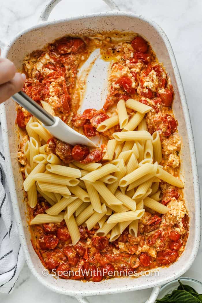 Baked feta pasta is a recipe that proves baked dishes can be both simple and amazing. Garlic, cherry tomatoes, and feta cheese are roasted in the oven until perfectly soft, then stirred into a creamy sauce that hugs every piece of pasta. This one-pan vegetarian pasta is both comforting and fresh, with bold roasted flavors and a creamy finish. It’s a baked pasta dish you’ll want to make again and again. #spendwithpennies adding pasta to tomatoes and feta in dish to make Baked Feta Pasta