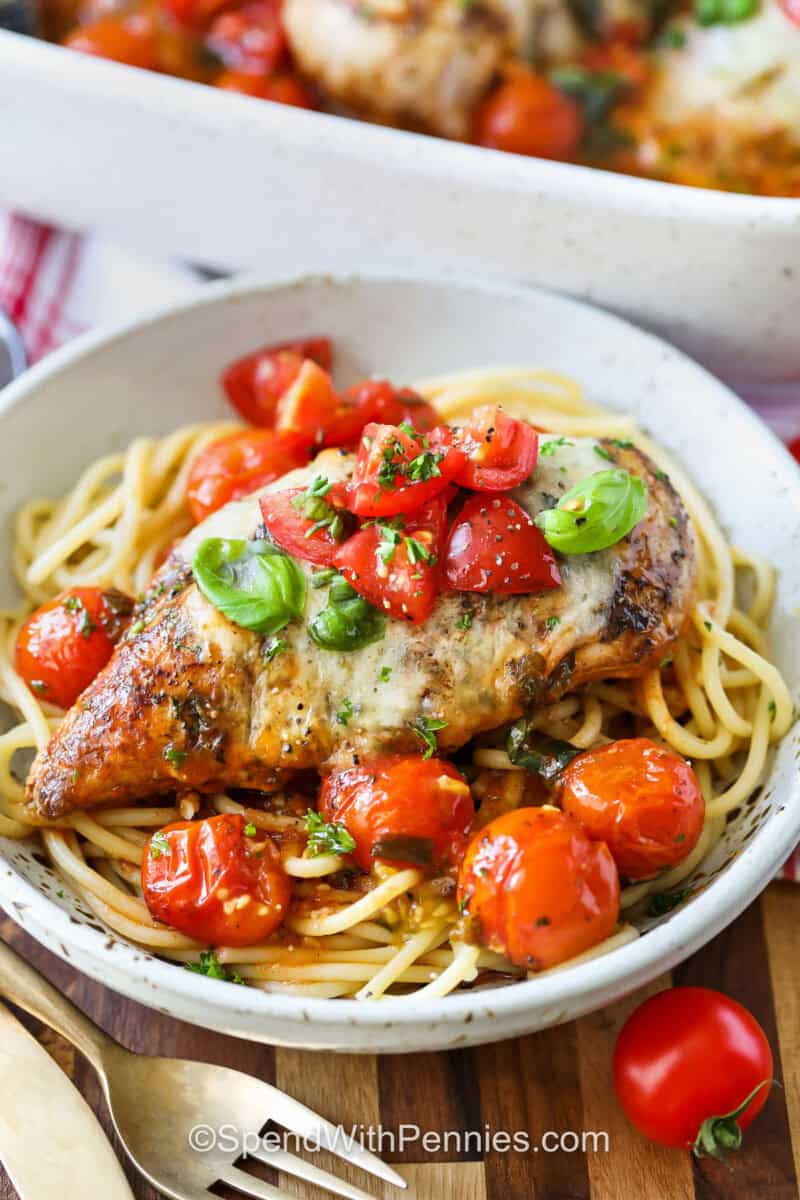 Bruschetta chicken is an easy baked recipe that delivers bold Italian flavors. Juicy chicken breast is marinated in balsamic, nestled in marinara, and baked with mozzarella and cherry tomatoes. Topped with a fresh tomato basil mixture, it’s a meal that’s perfect for serving with pasta, roasted vegetables, or a crisp salad. #spendwithpennies bowl of Bruschetta Chicken with tomatoes on spaghetti
