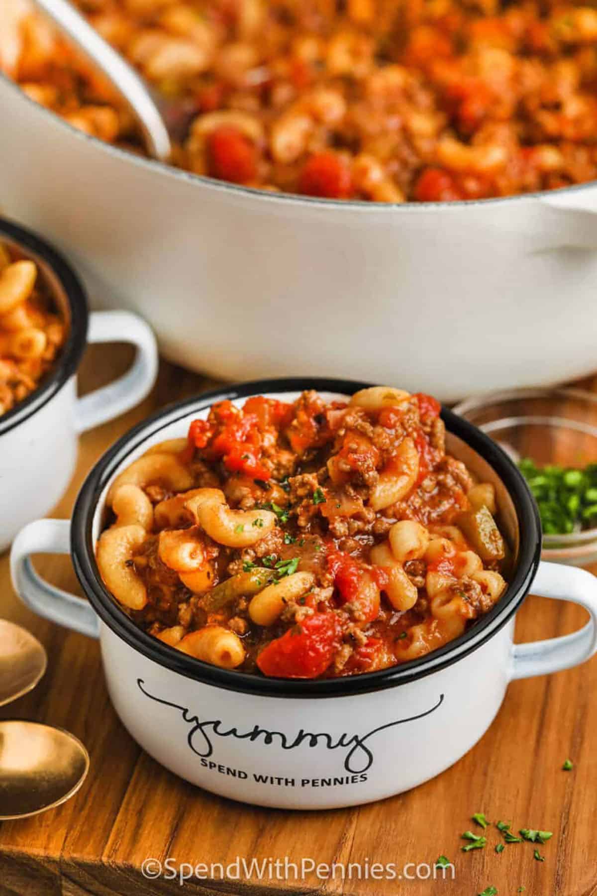 This hearty goulash recipe is the perfect one-pot meal for busy weeknights! Made with lean ground beef, tender elbow macaroni, and a rich tomato-based sauce, it comes together in under 30 minutes. Add a sprinkle of cheddar or mozzarella cheese on top for a comforting finish. Simple ingredients, bold flavor, and easy cleanup make this a go-to dinner your whole family will love. #spendwithpennies dish of Grandma's Goulash with a pot full in the back