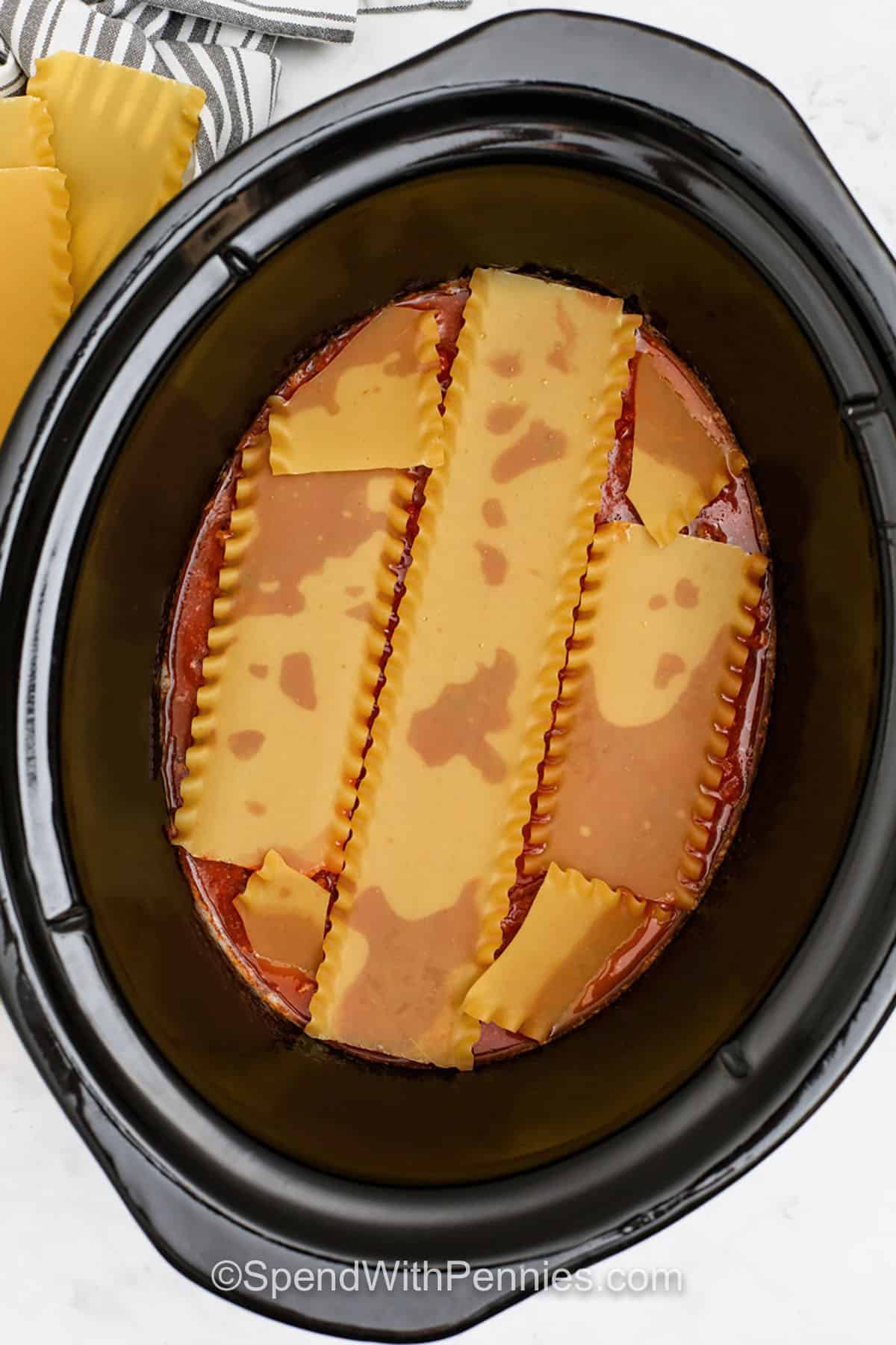 This crockpot lasagna recipe has everything you love about classic lasagna made easy in the slow cooker. Tender pasta, rich ricotta, seasoned beef, and plenty of melted cheese create layers of comfort and flavor. Each bite is warm, hearty, and satisfying. Serve it with garlic bread or a fresh green salad for a cozy meal everyone will enjoy. #spendwithpennies adding lasagna noodles layer to pot to make Crockpot Lasagna