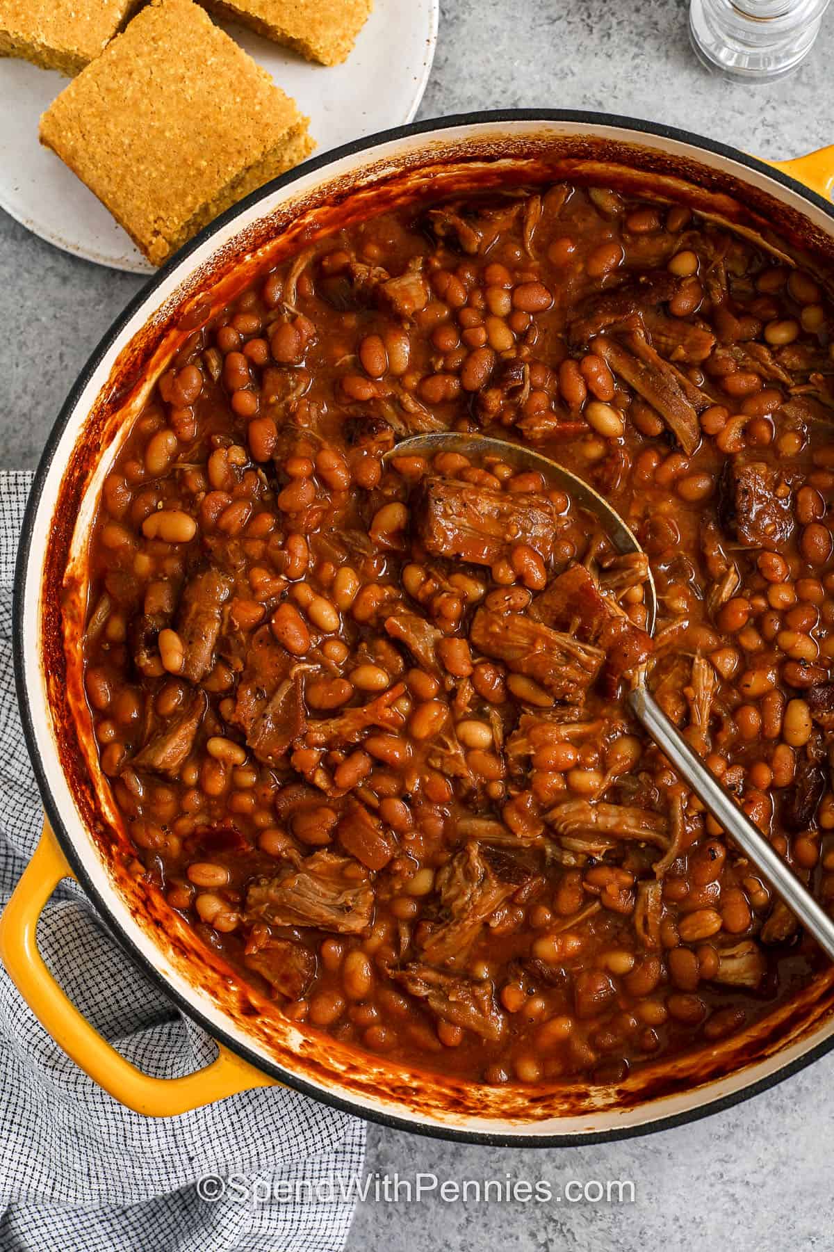 Pork and beans is a comfort food recipe where tender shredded pork meets saucy beans in a sweet, smoky broth. Baked beans, white beans, and savory pork are cooked together with bacon, onion, garlic, and spice for a hearty meal that tastes just as good reheated. Serve with coleslaw, bread, or rice for a dish that’s filling and delicious. This one always earns a spot at the table. #spendwithpennies Pork and Beans in a pot with plate of corn bread