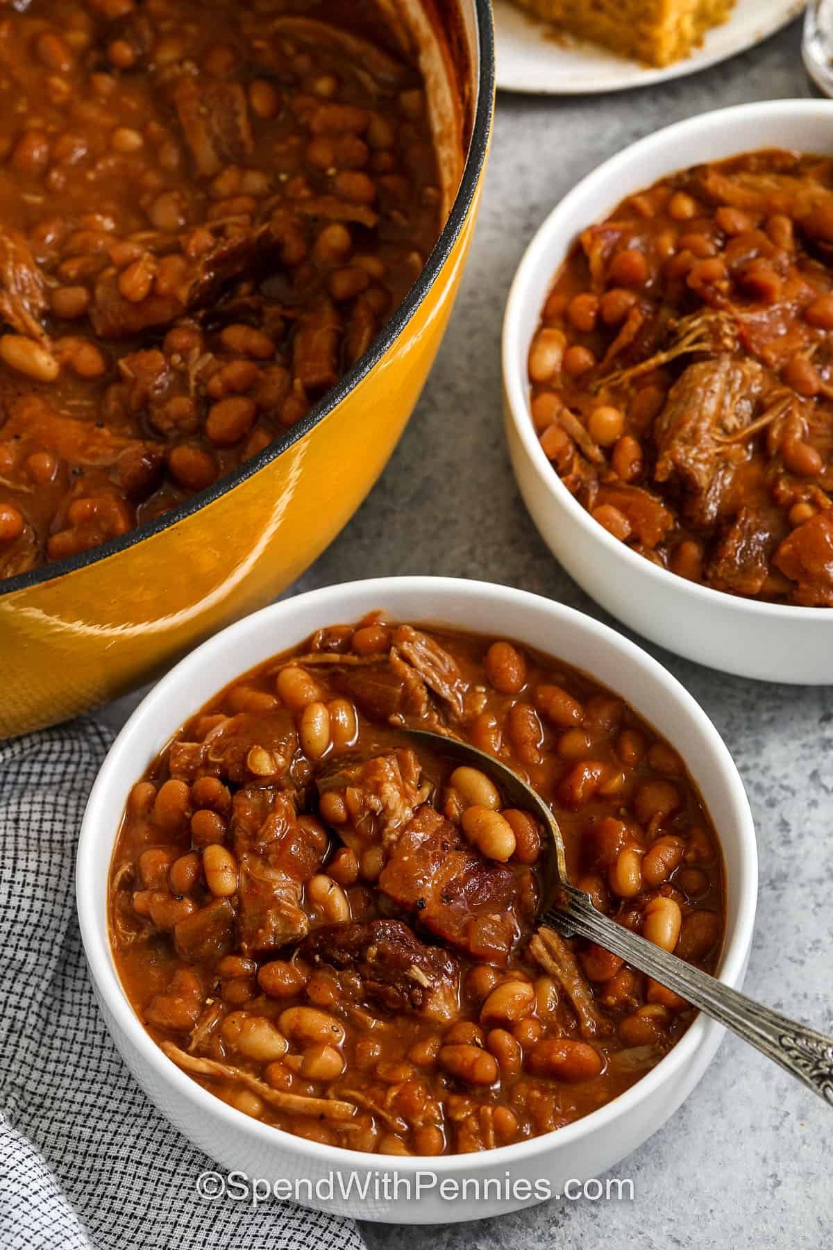 This pork and beans recipe combines tender pork shoulder, beans, and a tangy sauce in one hearty dish. Bacon, onion, and garlic give savory depth, while molasses, vinegar, and mustard add that sweet and zesty flavor. This recipe makes a generous pot perfect for feeding a crowd or enjoying as leftovers. Serve with rice, cornbread, or fresh coleslaw for a full meal everyone will love. #spendwithpennies bowls of Pork and Beans with pot full